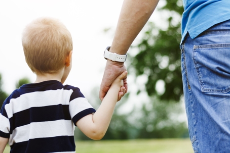 A father walking with his young child while holding the child's hand.