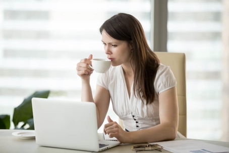 A woman sips coffee from a mug while working on a laptop.