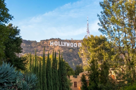 The Hollywood sign seen through a bunch of trees with blue skies and clouds overhead.